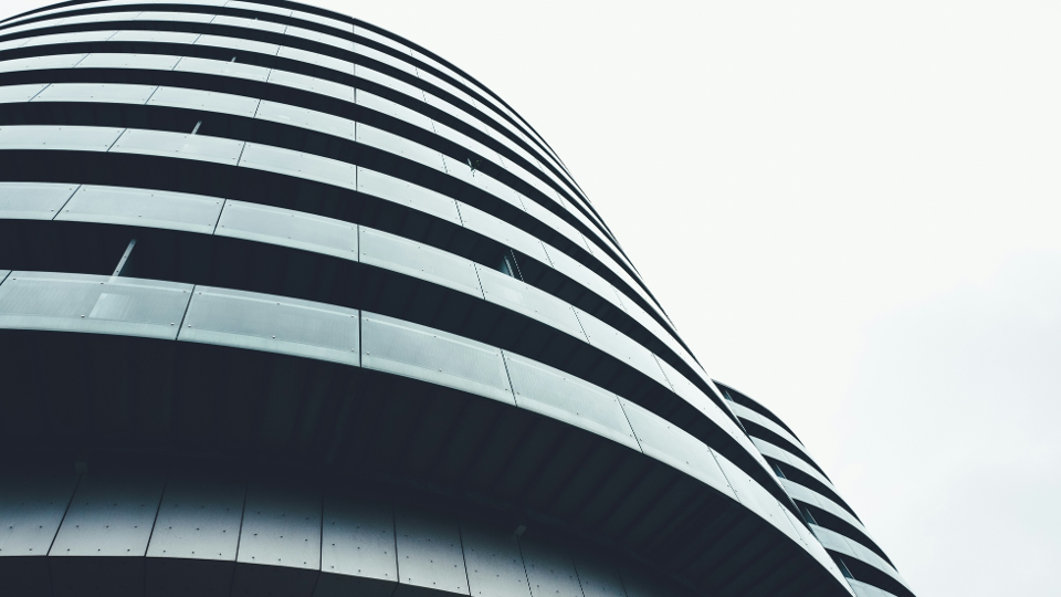 Low-angle view of a modern, curved building with horizontal black and glass balcony lines, set against a pale, overexposed sky. The architecture creates a sleek, futuristic, and minimalist aesthetic.